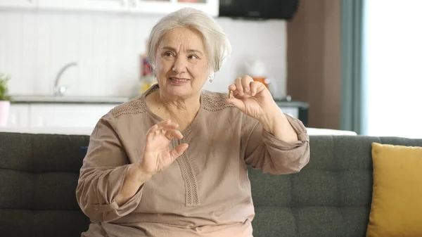 An elderly white-haired woman points to a pill on the living room sofa. She points out that the medicine in her hand is perfect. Happy old woman showing health care, disease treatment, vitamins.