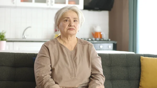 stock image Thoughtful old woman in her home or nursing home. Health services for the elderly. Portrait of elderly woman looking at camera while sitting on sofa in living room.