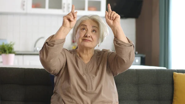 The old woman points to the ad space above. Look here. Beautiful funny happy old woman showing copy space above head while sitting on sofa at home looking up to show message.
