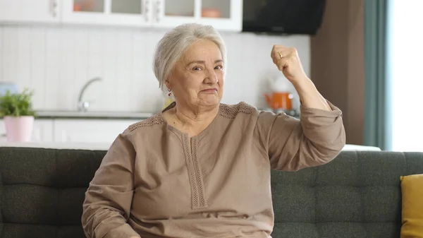 An elderly woman raises her arms and shows biceps to the camera at her home. Feminism and women\'s power. The old woman states that she is still strong. Women, Healthy life, women\'s health concepts.