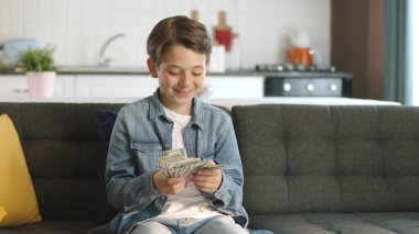 Beautiful little boy very happy with his money.Winning the online lottery lottery. The boy is making a fan out of dollars. Excited little boy enjoying his dollar at home alone. clipart