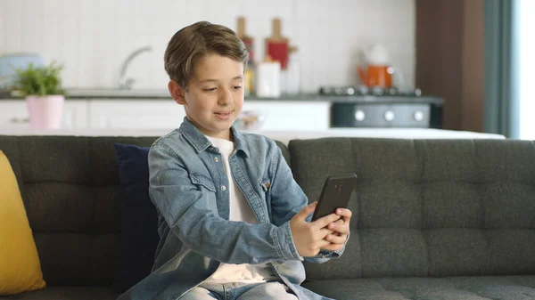 Friendly little boy making a video call on his cell phone. Little funny boy making online video call with friends on smartphone while sitting on sofa at home.