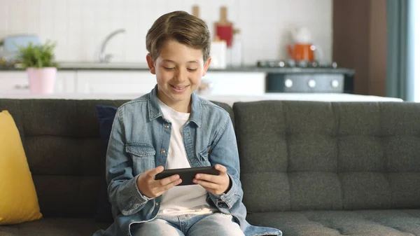 Boy using phone to play games. Little funny boy playing smartphone game while sitting on sofa at home, next generation games, happy childhood.