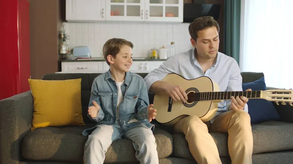 Father playing guitar to his little son while sitting on sofa at home. Young father having fun singing songs with his son and spending happy time together. Portrait of happy father and son.