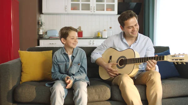 Father playing guitar to his little son while sitting on sofa at home. Young father having fun singing songs with his son and spending happy time together. Portrait of happy father and son.
