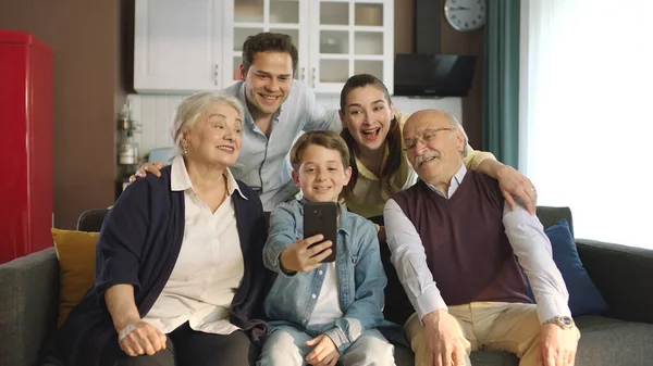 Young couple with children, their son and elderly parents sitting on sofa in living room, taking self portraits together. Portrait of happy cheerful big family smiling at camera in cozy living room.