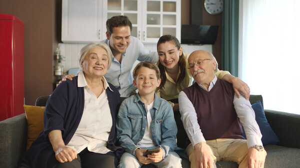 Young couple with children, their son and elderly parents sitting on sofa in living room, taking self portraits together. Portrait of happy cheerful big family smiling at camera in cozy living room.