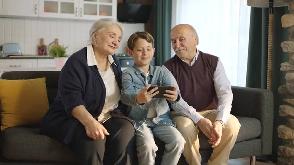 Little boy visiting his grandparents. Happy elderly couple sitting on sofa and chatting with their little grandchild. They are taking selfies together.
