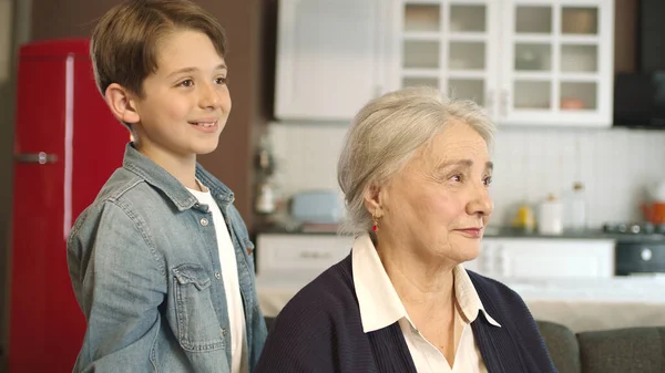 The child pays a visit to his grandmother, who is sitting in his armchair. Portrait of happy grandmother and granddaughter looking at empty advertising space to the right of the screen.