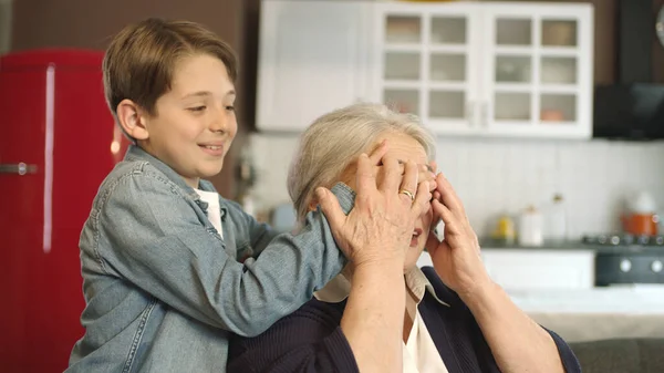 The child approaches his grandmother, who is sitting in his armchair, and closes his eyes. The boy pays his grandmother a surprise visit. They hug each other. Portrait of happy grandmother and grandch