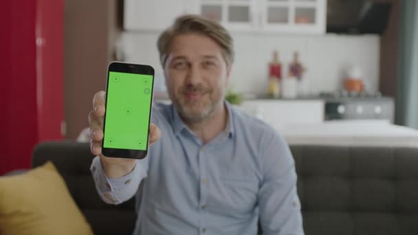 若い男が自宅で快適なアームチェアに座って カメラで緑の点でスマートフォンを指しています 広告のためのクロマキーグリーンスクリーンスマートフォンのセット — ストック動画
