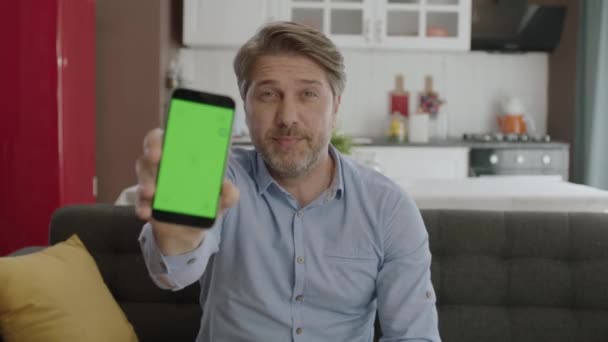 若い男が自宅で快適なアームチェアに座って カメラで緑の点でスマートフォンを指しています 広告のためのクロマキーグリーンスクリーンスマートフォンのセット — ストック動画