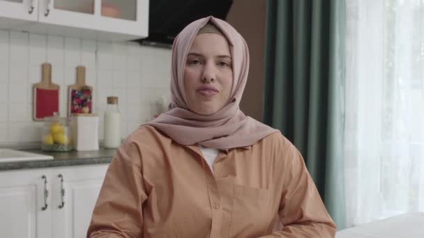 キッチンに立ってカメラを見て服を着たヒジャーブの若い大人のイスラム教徒の女性の笑顔を自信を持って 若い大人の女性がカメラを見て微笑んだ ムスリム女性の肖像画 — ストック動画