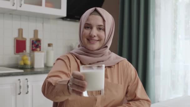 ヒジャーブ州の女性は カメラに牛乳のガラスを与えます 笑顔若いです女性でヒジャーブでテーブルでキッチンと与えミルクにカメラ — ストック動画