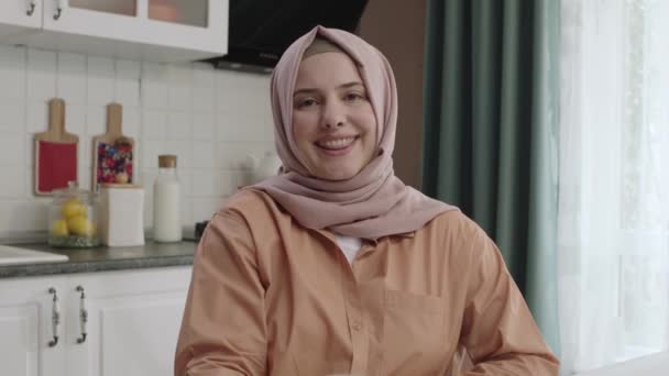 キッチンに立ってカメラを見て服を着たヒジャーブの若い大人のイスラム教徒の女性の笑顔を自信を持って 若い大人の女性がカメラを見て微笑んだ ムスリム女性の肖像画 — ストック動画