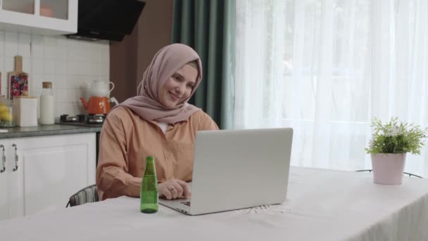 ヒジャブの服を着た女性は台所でラップトップで作業し フリーランスのリモートビジネスメッセージングメッセージは 休憩を取り 自然のミネラルウォーターを飲み 健康管理の一口を取り 脱水を防ぎます 日常生活 — ストック動画