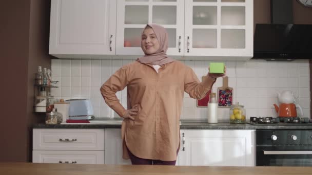 キッチンで何か緑を持っているヒジャーブの女性は 製品を示し 笑顔と陽気で想像上のオブジェクトを提示します クリエイティブ3Dアーティストは 緑のボックスを任意の製品に置き換えることができます — ストック動画