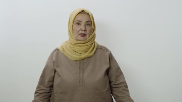 白い背景に孤立したスタジオで高齢者のヒジャーブの女性 白い背景に2本の指で画面の下部にスカーフポイントを身に着けている高齢者のイスラム教徒の女性 — ストック動画