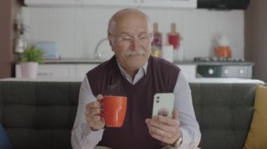 Mutlu 70 'ler erkek kullanıcısı kahve içerken internetten video çekiyor, kanepede oturup selfie çekiyor, yaşlı büyükbaba modern teknolojiyi kullanmayı öğreniyor..