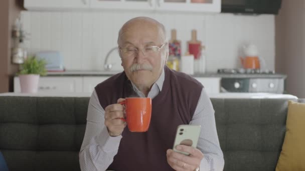 老人は自宅でコーヒーを飲みながらスマホで時間を過ごす 男は毛深い携帯電話で見たSmsやビデオを見ている 高齢者における技術利用の概念 — ストック動画
