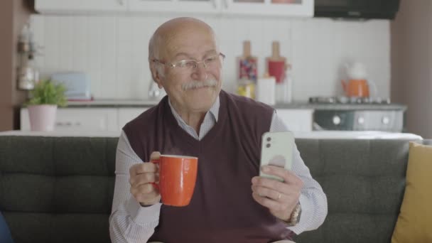 老人は電話で番号をダイヤルし 老人は自宅でコーヒーを飲みながら携帯電話にSmsを送信します 80歳の男性はスマートフォンでモバイルアプリやSmsやニュースを見て — ストック動画