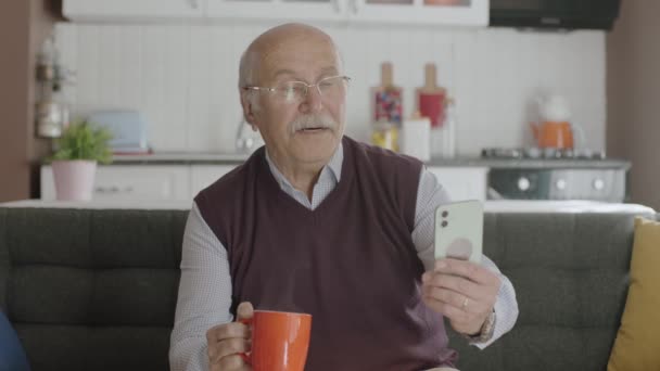 自宅でコーヒーを飲みながらスマホで面白い動画を見ている老人は スマホで見たSmsや動画を笑いながらカメラを見ている — ストック動画