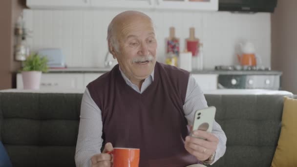 自宅でコーヒーを飲みながらスマホで面白い動画を見ている老人は スマホで見たSmsや動画を笑いながらカメラを見ている — ストック動画