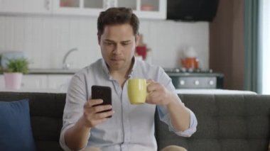 Akıllı telefon kullanan ve evde kahve içen düşünceli genç bir adam. Sosyal medya hesaplarını akıllı telefondan tararken kahve içen genç kullanıcı gördüğü haberlere ya da mesajlara şaşırıyor..