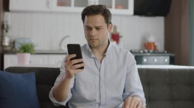 Sıradan giyimli genç adam evdeki koltukta akıllı telefon kullanıyor. Genç adam mobil teknoloji kullanıcısı dijital uygulamalar üzerinde çalışıyor, internette bilgi arıyor, evde veya ofiste mesaj yazıyor..
