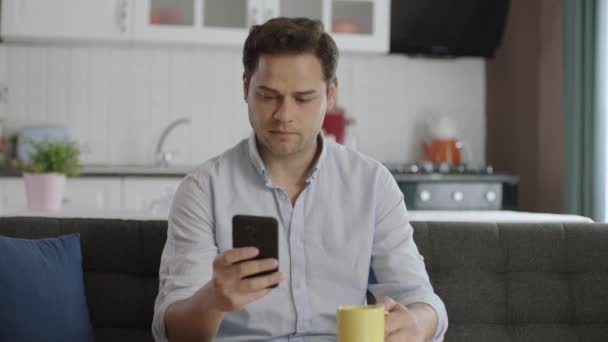 Leende Ung Man Som Använder Smartphone Och Dricker Kaffe Hemma — Stockvideo