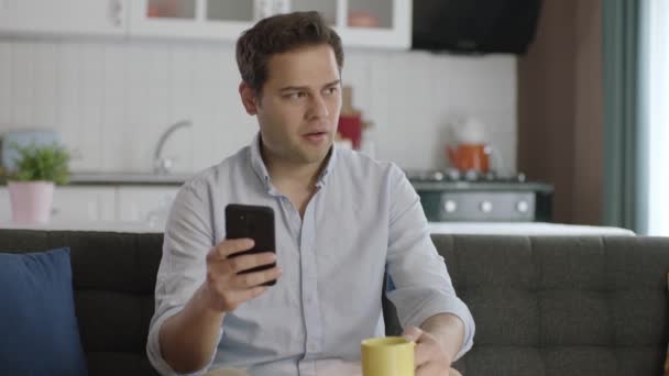 Tänksam Ung Man Som Använder Smartphone Och Dricker Kaffe Hemma — Stockvideo