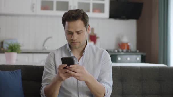 自宅でソファの上でスマートフォンを使用してカジュアル服装の若い男 デジタルアプリに取り組んで若い男のモバイル技術ユーザー オンライン情報を検索 自宅やオフィスでテキストメッセージ — ストック動画