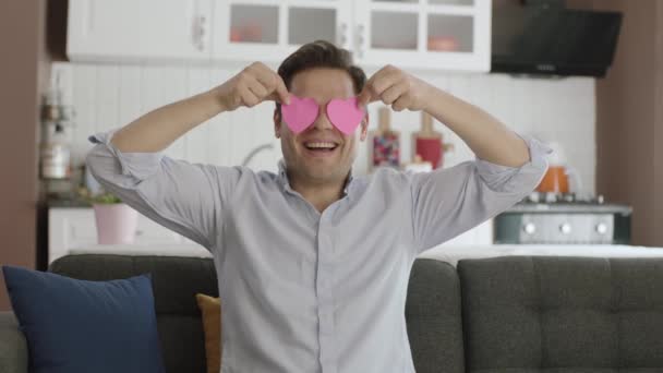2枚の紙の心で遊んでいる幸せな男のビデオ 赤いハートの紙で作られた彼の家のソファで踊る男 バレンタインデー目の愛ロマンチックなコンセプト — ストック動画