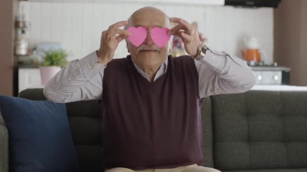 2枚の紙の心で遊んでいる幸せな老人のビデオ 老人は赤い紙の心で彼の家のソファで踊っています バレンタインデー目の愛ロマンチックなコンセプト — ストック動画