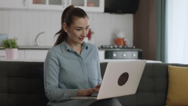 自宅のソファでノートパソコンで働く若い女性 新しい技術の使用 ラップトップ上の幸せなビジネス女性のフリーランサー 技術デバイスとの時間を過ごす 指でコンピュータに広告スペースを表示します — ストック動画