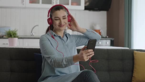 自宅で音楽を聴くためにワイヤレスヘッドフォンを使用してソファに座っている女性 彼女は彼女の平和な家庭で彼女の携帯電話に接続されたヘッドフォンで聞いている音楽に踊っている幸せな女性 — ストック動画