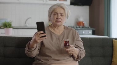Cep telefonu tutan mutlu yaşlı bir büyükanne, çay içerken sevdikleriyle görüntülü konuşma. Modern teknoloji aygıtı kullanmayı öğrenen yaşlı bir kadın. Yaşlılarda teknoloji kullanımı.