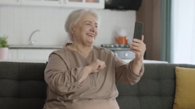 Modern teknoloji aletini kullanmayı öğrenen yaşlı bir kadın. Cep telefonu tutan mutlu yaşlı bir büyükanne, sevdikleriyle video görüşmesi, el sallayan mutlu yaşlı bir büyükanne. Yaşlılarda teknoloji kullanımı.