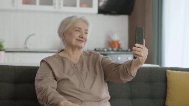 Yaşlı kadın modern teknoloji aletlerini kullanmayı öğreniyor. Mutlu yaşlı büyükanne elinde cep telefonuyla cep telefonuyla konuşuyor, selfie çekiyor, evdeki kanepede dinleniyor. Yaşlılarda teknoloji kullanımı.