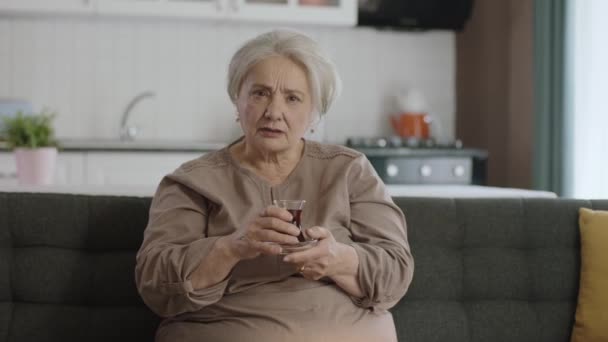 快乐的老妇人一边喝茶一边在电视上看一部有趣的电影 老妇人独自坐在沙发上看一部神秘的电视电影 老人类 — 图库视频影像