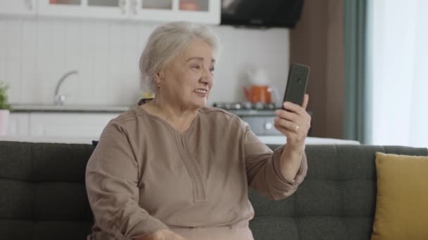 幸せな老祖母の携帯電話を保持し 愛する人とのビデオ通話を行うと 幸せな老祖母手を振る 高齢女性は現代の技術デバイスを使用することを学ぶ 高齢者における技術利用 — ストック動画