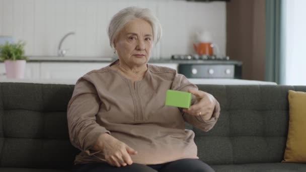 老妇人在扶手椅上拿着绿色的东西 展示着一种产品 微笑着 呈现着一个快乐的 想象的物体 创意3D艺术家可以用他们想要的任何产品替换绿色盒子 — 图库视频影像