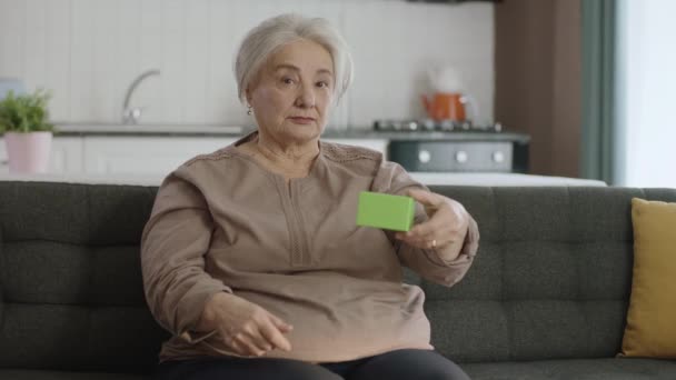 老妇人在扶手椅上拿着绿色的东西 展示着一种产品 微笑着 呈现着一个快乐的 想象的物体 创意3D艺术家可以用他们想要的任何产品替换绿色盒子 — 图库视频影像