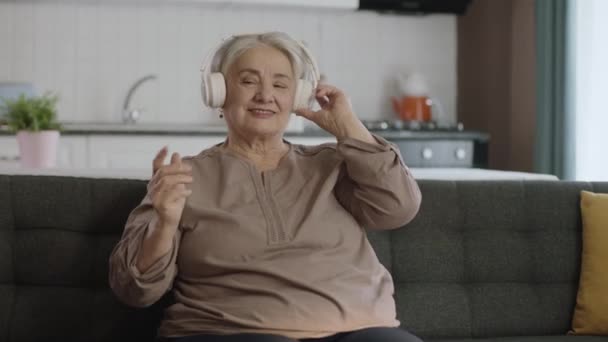 引退後の人生を楽しむ熟女陽気な女性 ソファの上にヘッドフォンで音楽を聴いている老人のカラフルで面白い瞬間 面白いです古いです女性とともにヘッドフォン歌と踊り — ストック動画