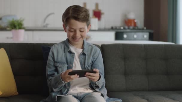 男の子はゲームをプレイするために電話を使用しています 自宅でソファに座っている間に小さな面白い男の子はスマートフォンゲームをプレイし 新世代のゲーム 幸せな子供時代 — ストック動画