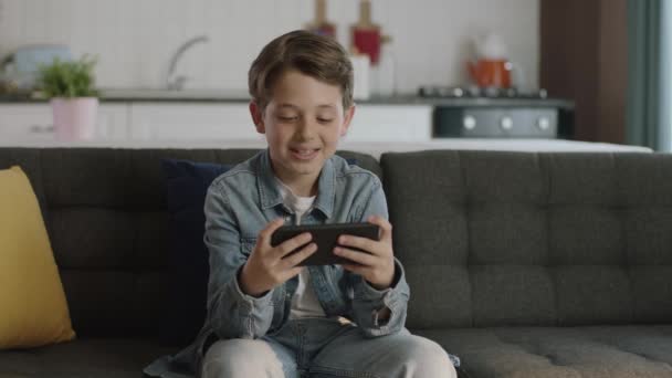 男の子はゲームをプレイするために電話を使用しています 自宅でソファに座っている間に小さな面白い男の子はスマートフォンゲームをプレイし 新世代のゲーム 幸せな子供時代 — ストック動画