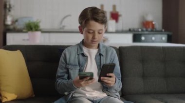 Teknoloji bağımlısı küçük çocuk. Meraklı çocuk kanepeye oturup iki akıllı telefonla oyun oynuyor. Evde internette sörf yapan küçük çocuk..