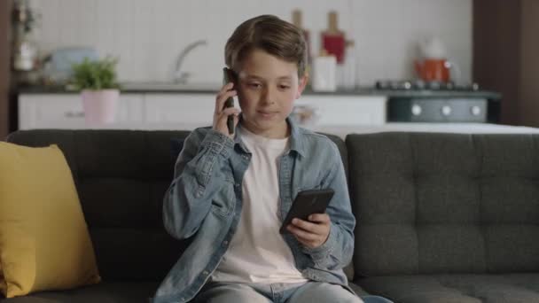 一个好奇的男孩坐在沙发上 用两台智能手机玩游戏 小男孩在家里上网 — 图库视频影像