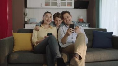 Modern, güzel, mutlu bir ailenin portresi. Genç ebeveynler ve küçük oğulları ellerinde akıllı telefonlarla kameraya gülümsüyorlar..