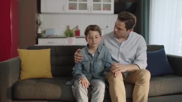 父は悲しい息子を慰めました 家でソファに座っている間 若い父と小さな息子の肖像画 父は学校や生活の中で問題を抱えている彼の若い息子を慰めます — ストック動画